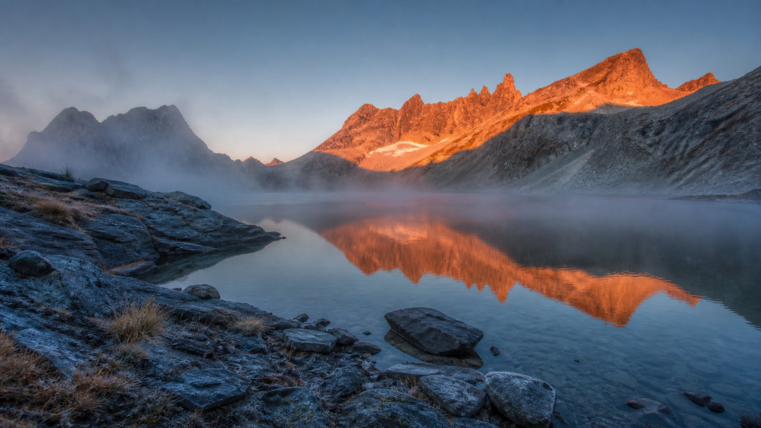 Small alpine lake reflecting a vivid warm ridge while the shore remains cool and blue, original 4K mountains wallpaper