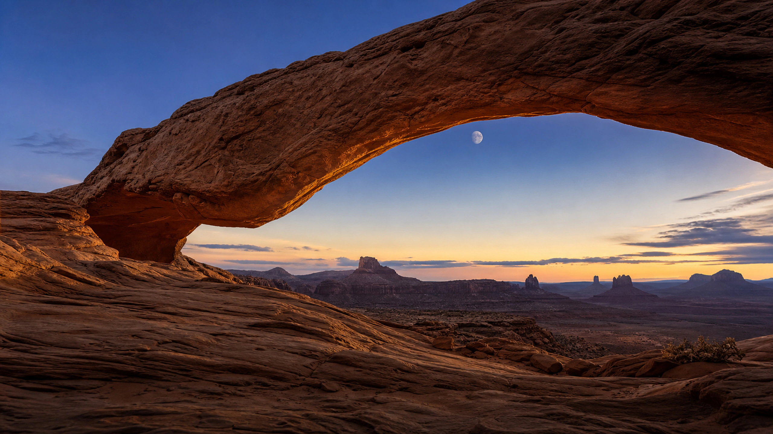 Early moon visible through a broad red-rock arch during blue-and-gold twilight, original 4K desert wallpaper