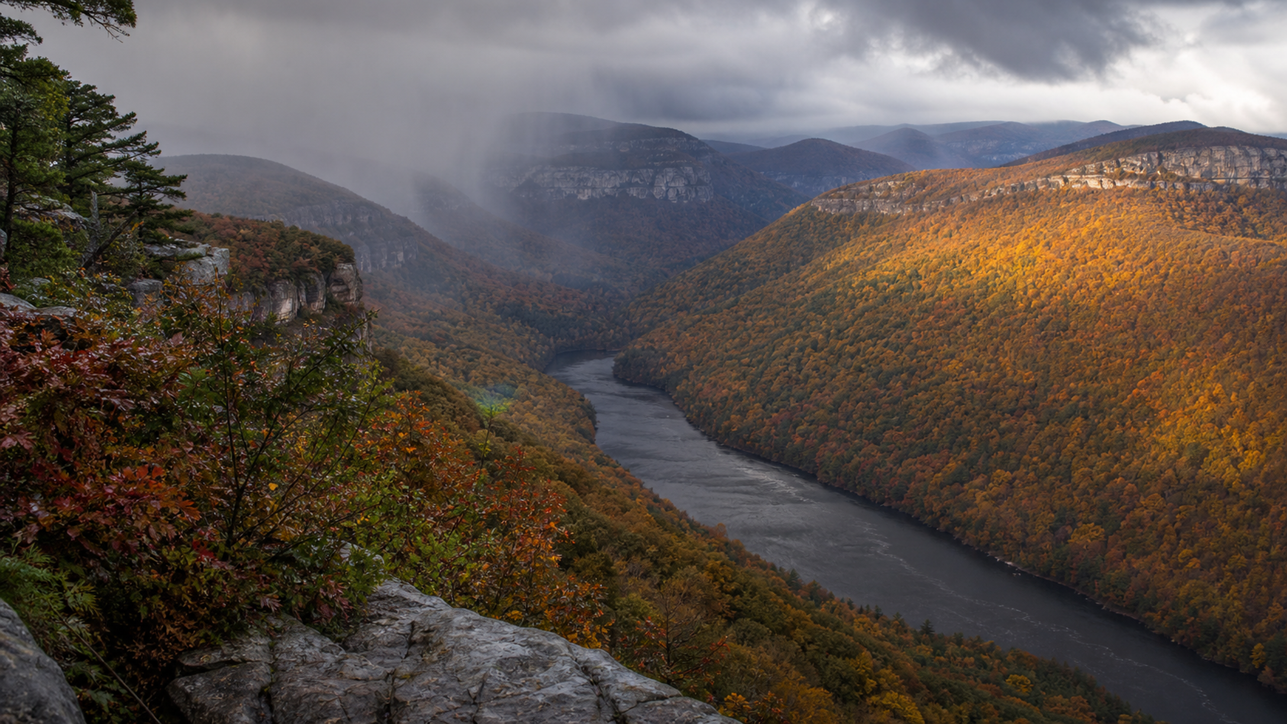 An original temperate karst valley covered in peak-autumn forest as one natural sunbreak crosses a copper slope beneath retreating rain curtains, original 4K seasons wallpaper