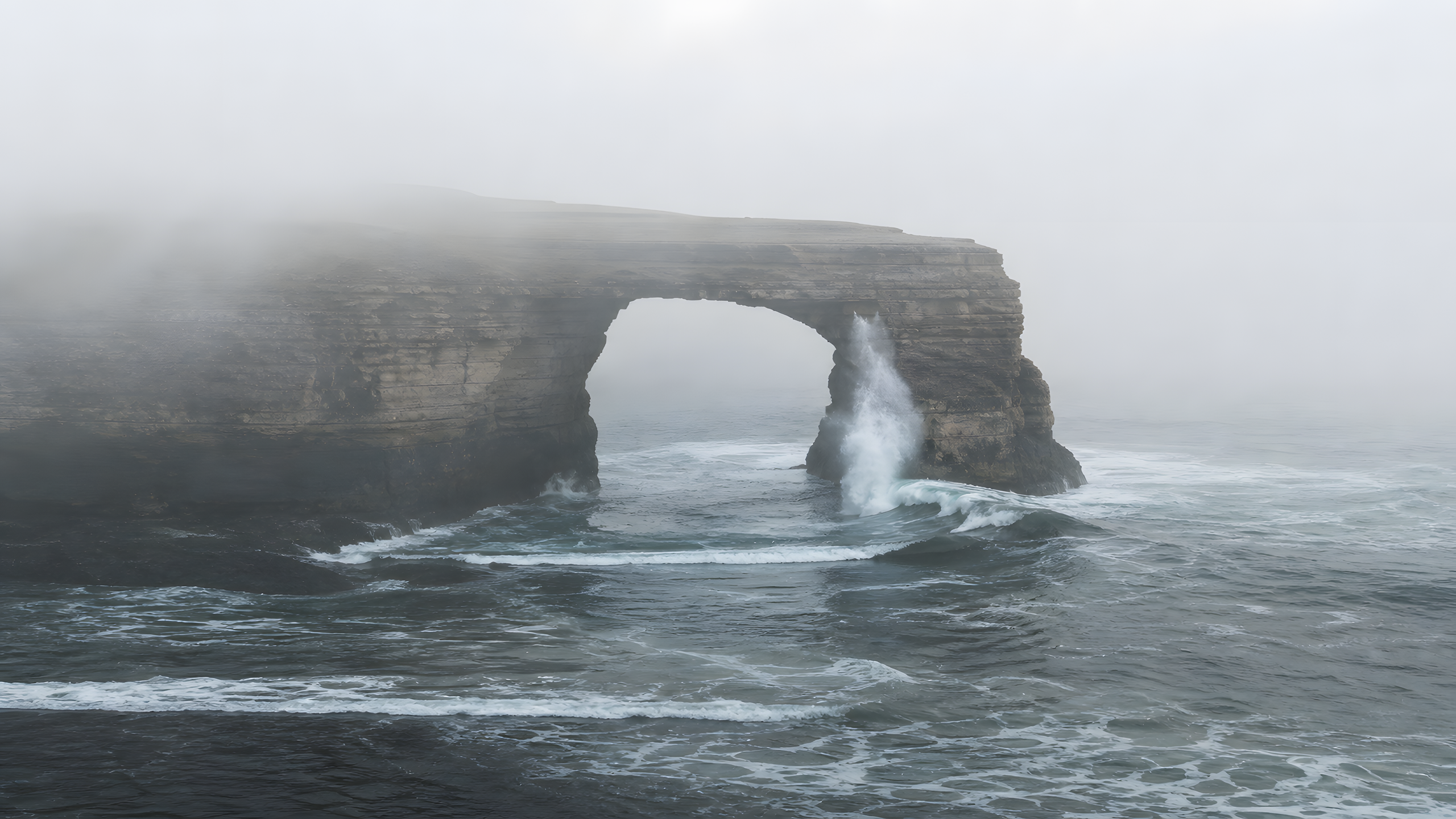 A broad swell passing through a pale sandstone sea arch and erupting against its far pillar in dense morning fog, original 4K ocean wallpaper