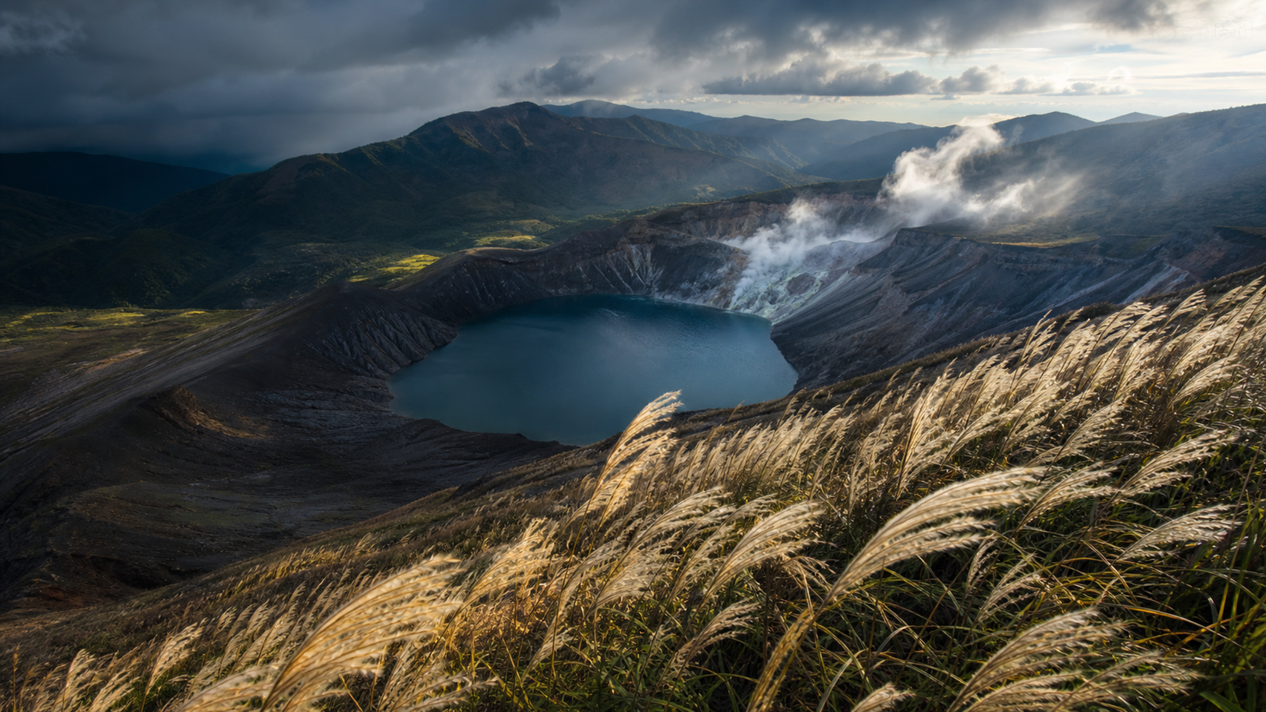 Broad silver-green grassland rolling across an ancient volcanic caldera, with one dark timber shelter beneath fast-moving afternoon cloud, original 4K travel wallpaper