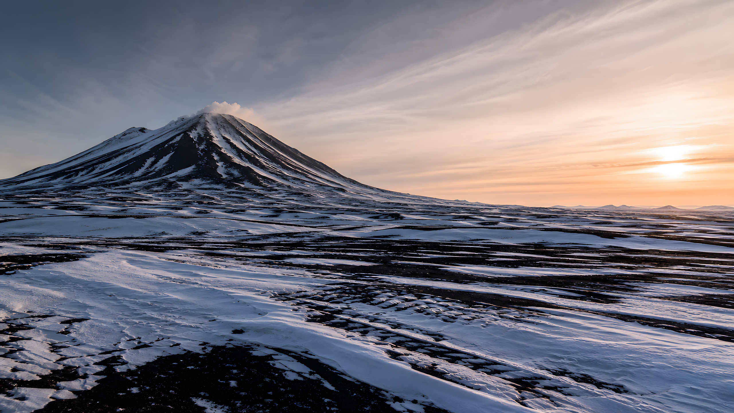 A solitary charcoal stratovolcano above a black plateau patterned by wind-sculpted snow ribbons, summit spindrift and pale apricot winter sunrise, original 4K mountains wallpaper