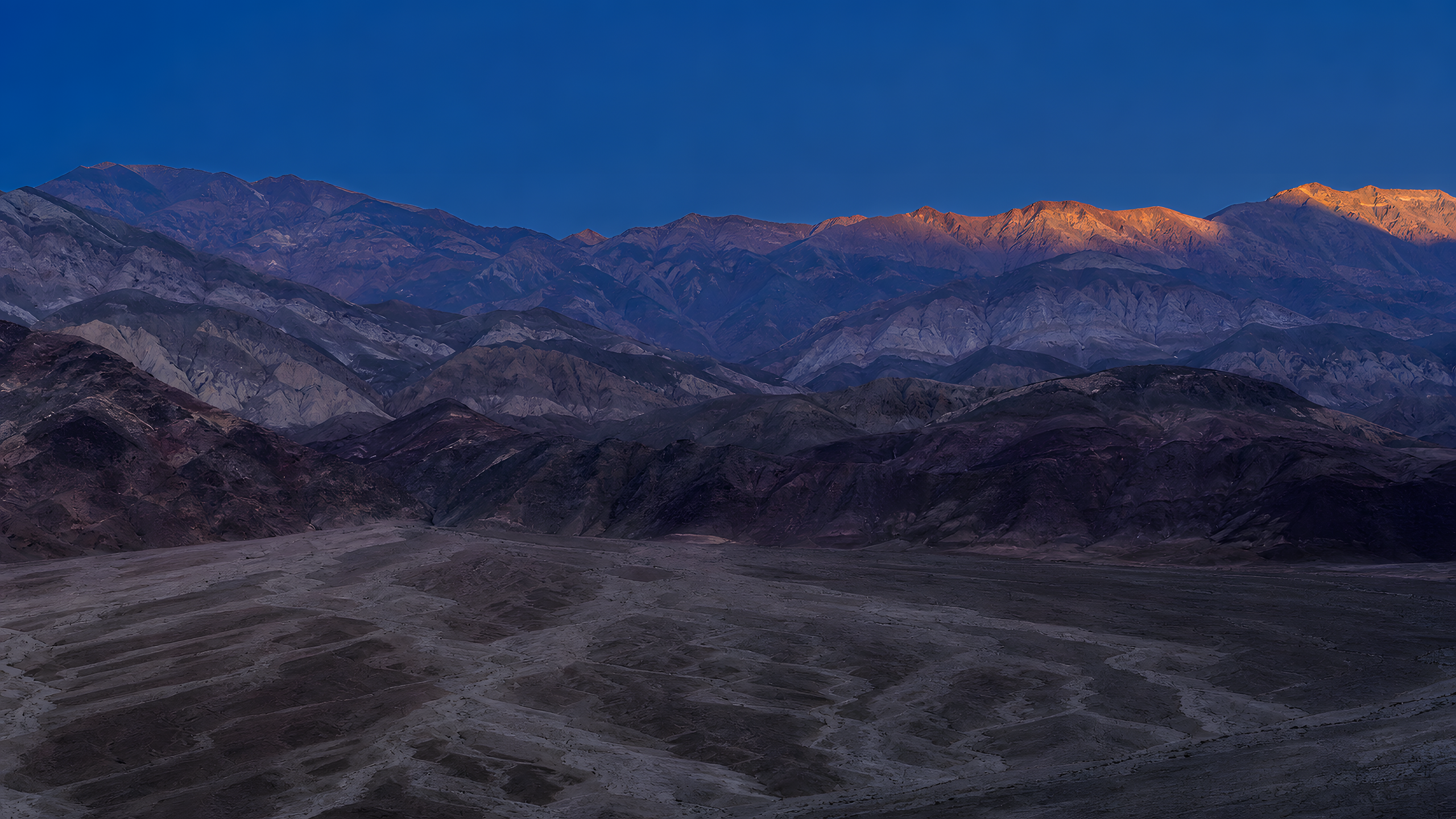 Layered arid fault-block ranges compressed by a long lens at blue hour, with ochre, mauve and charcoal mineral bands above branching alluvial washes, original 4K mountains wallpaper