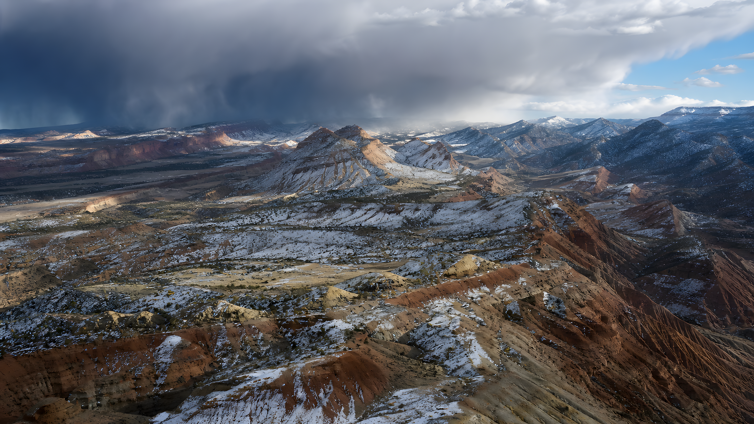 A high oblique view across red sandstone and pale shale mountains dusted by early-spring snow while a dark squall crosses the far basin and sunlight returns to foreground ridges, original 4K mountains wallpaper