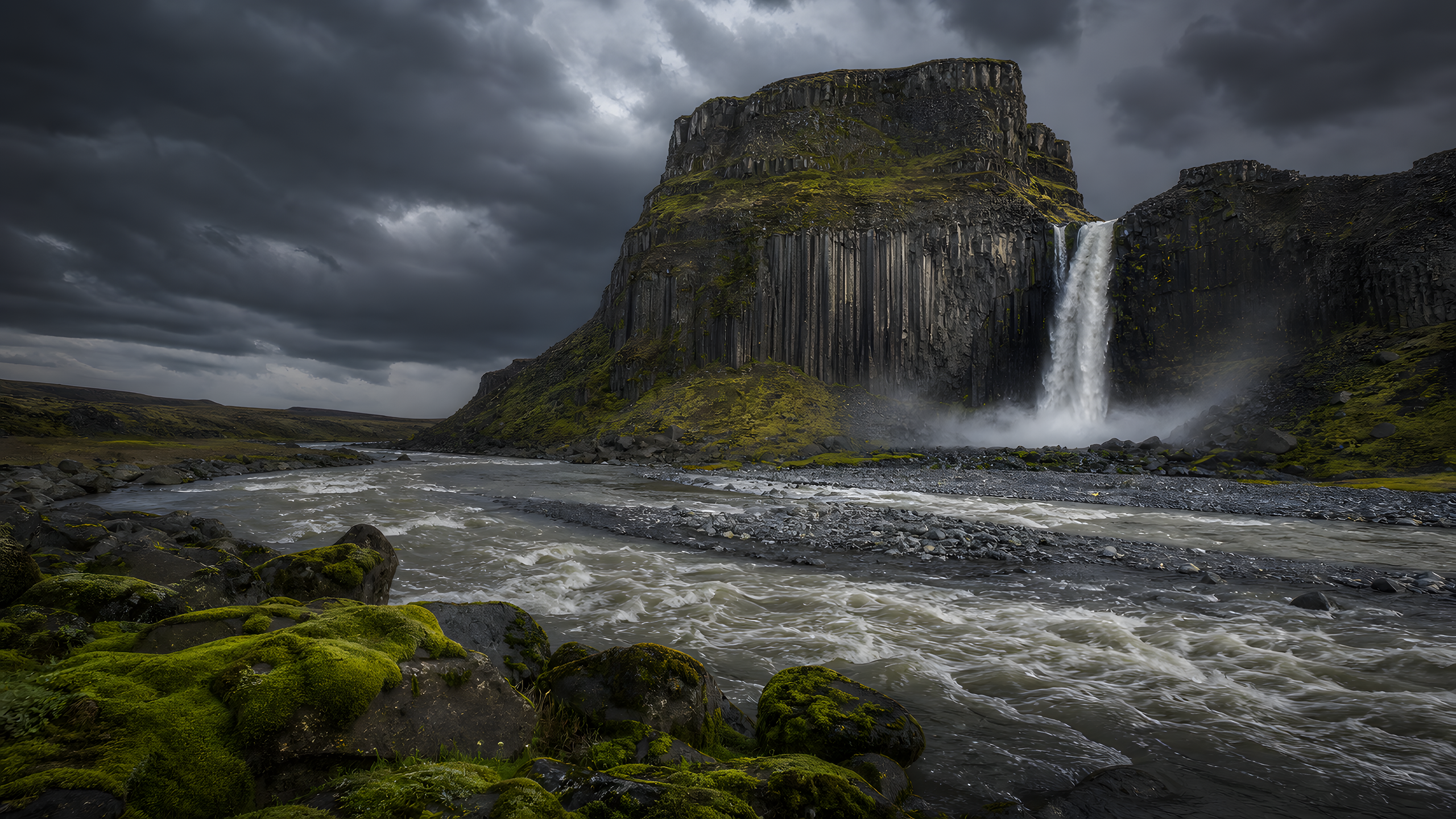 A rain-fed waterfall dropping from a columnar-basalt table mountain into a braided gray river beneath a charcoal storm front and cool moss-lit sunbreak, original 4K mountains wallpaper