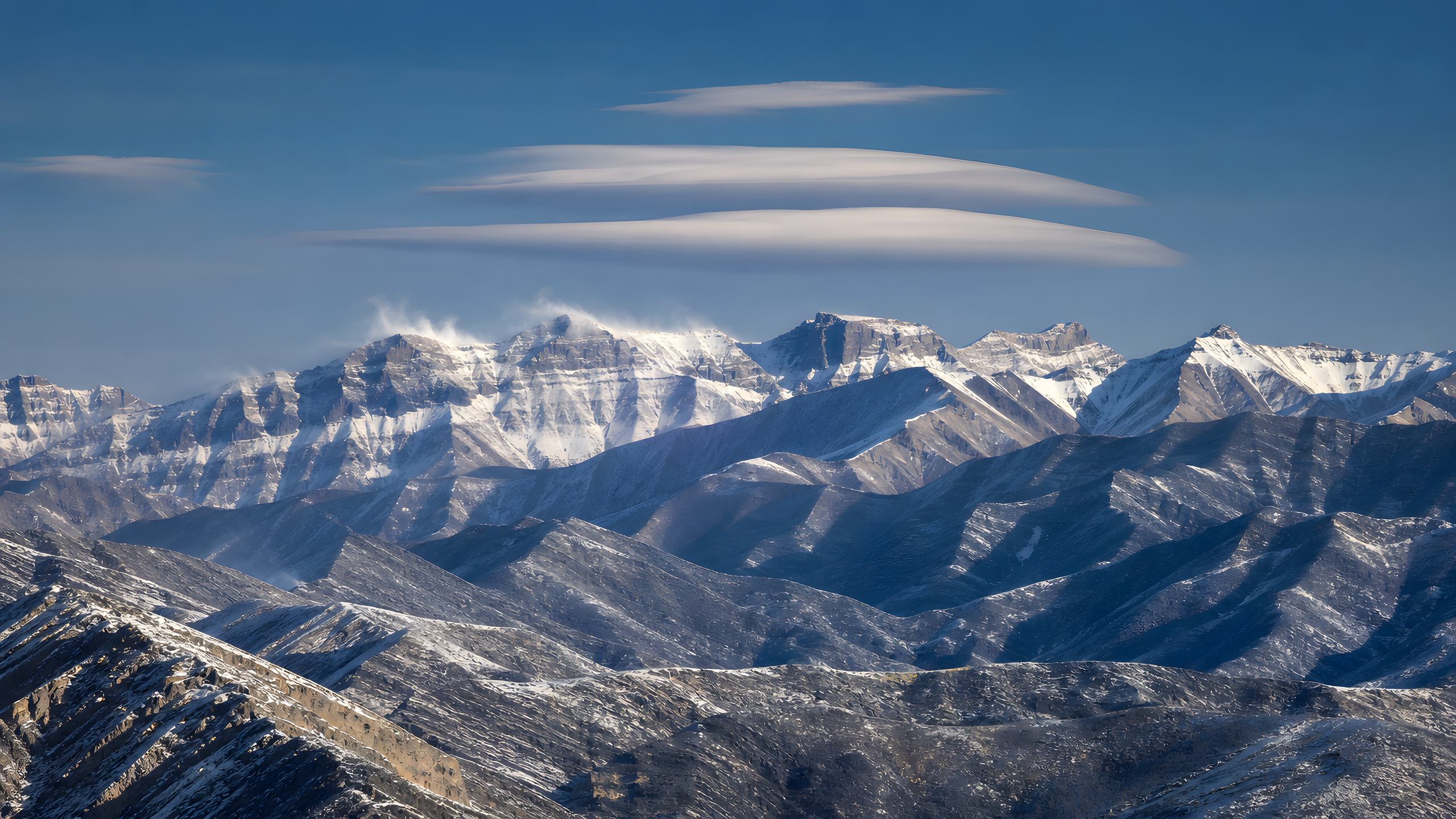 Complex overlapping winter ranges in alternating snowy and exposed slate bands beneath stacked lenticular clouds and wind-blown crest snow, original 4K mountains wallpaper