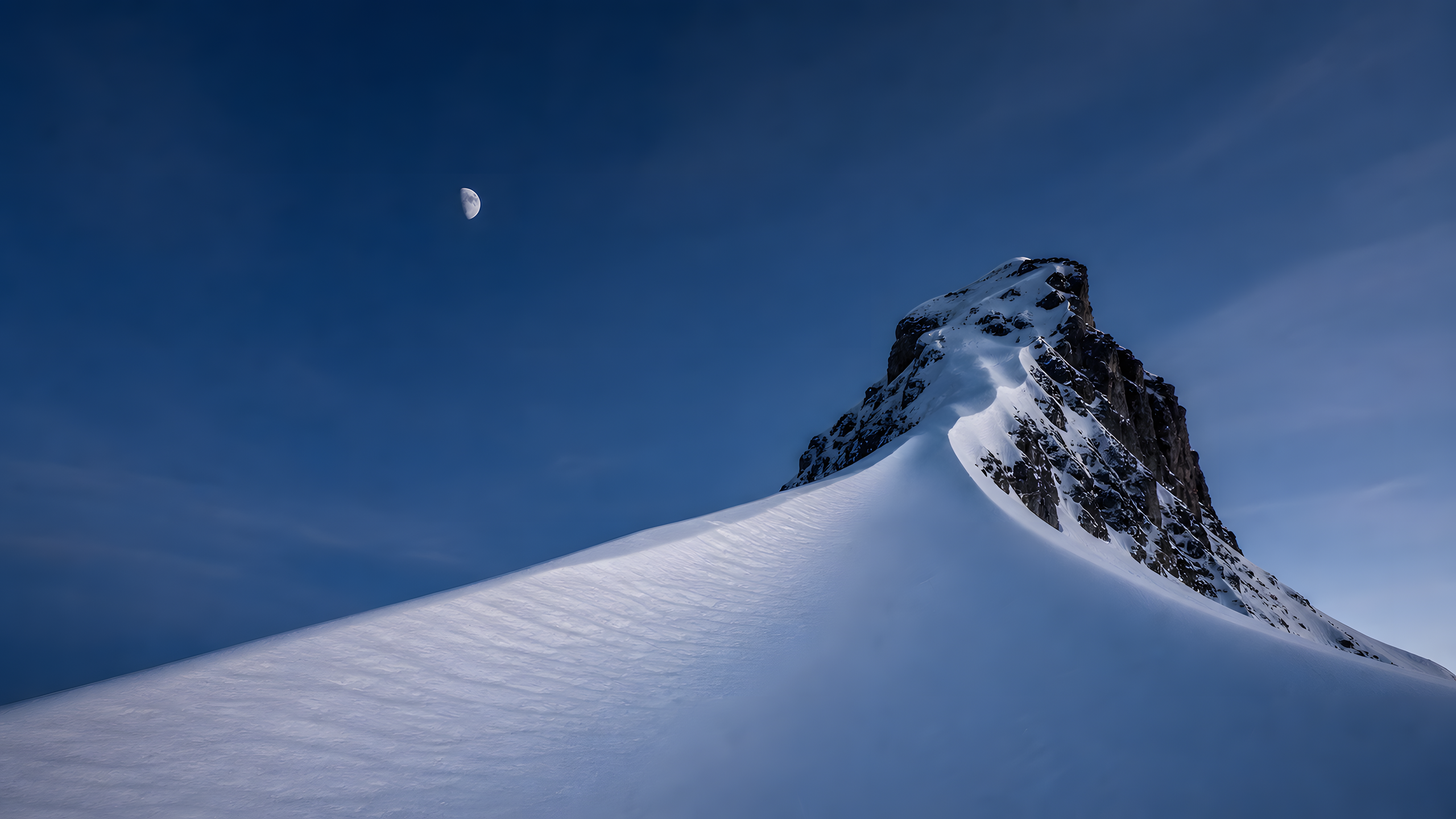 A minimalist predawn snow cornice rising diagonally toward a dark rock summit beneath a small waning moon and broad indigo negative space, original 4K mountains wallpaper