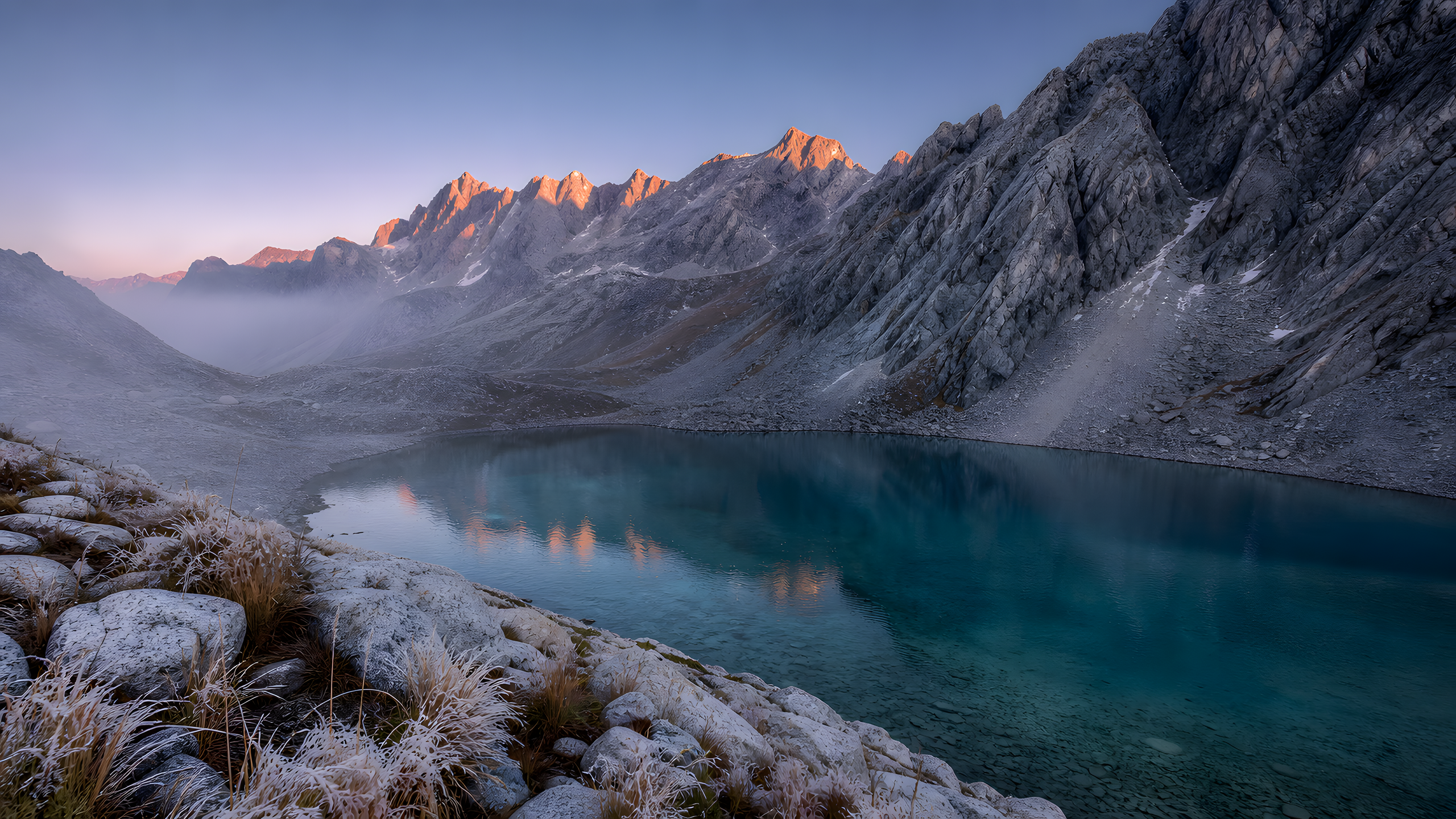 A cold turquoise glacial lake beneath rugged granite peaks touched by restrained blush alpenglow, seen from an elevated shoreline overlook with pale frost-tipped grasses and smooth foreground stones, original 4K pastel wallpaper