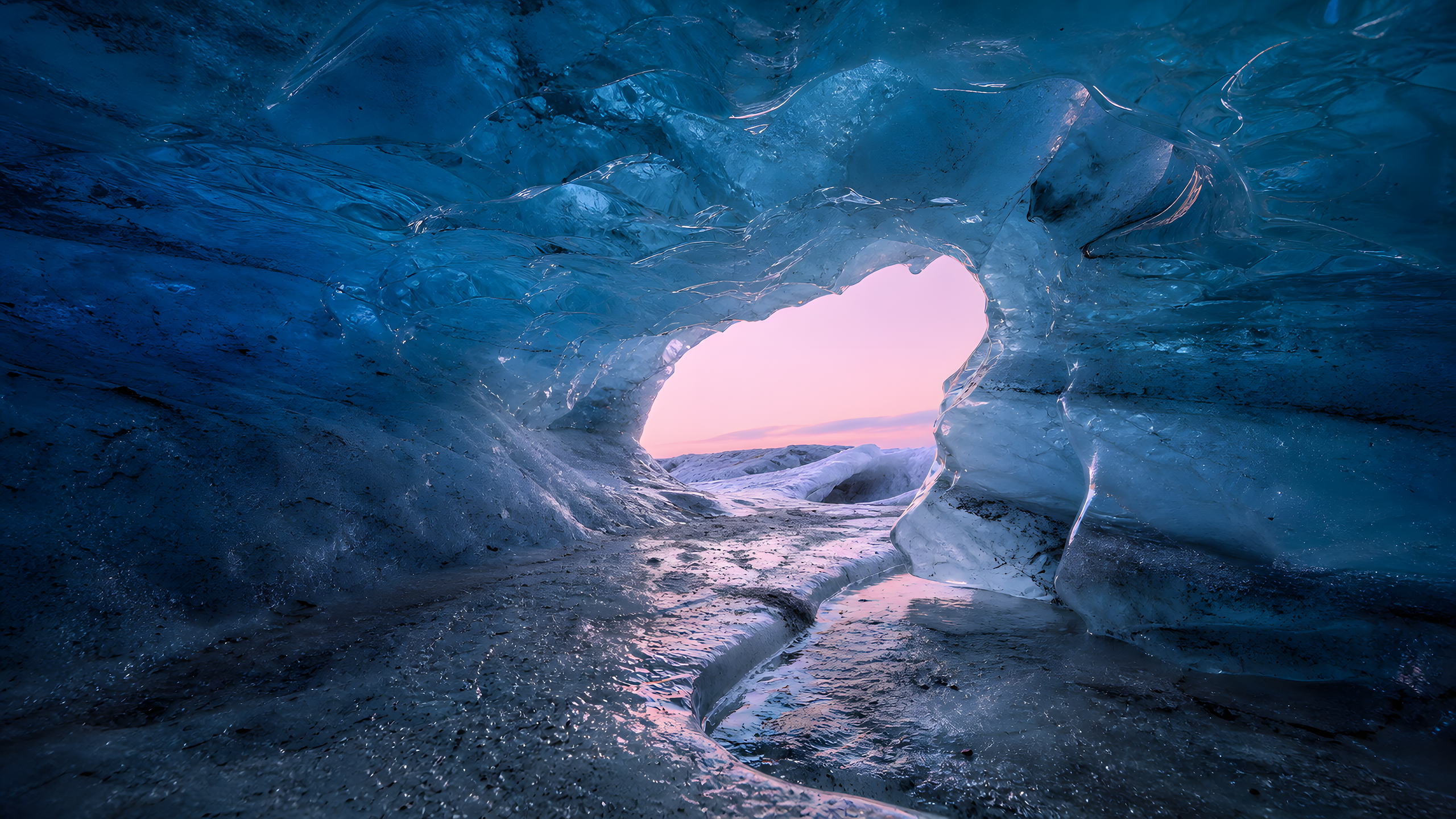 A naturally formed blue-white ice cavern with layered ice, trapped bubbles, mineral dust and melt channels, viewed low along a sinuous wet groove toward a pale pink winter-dusk opening, original 4K pastel wallpaper