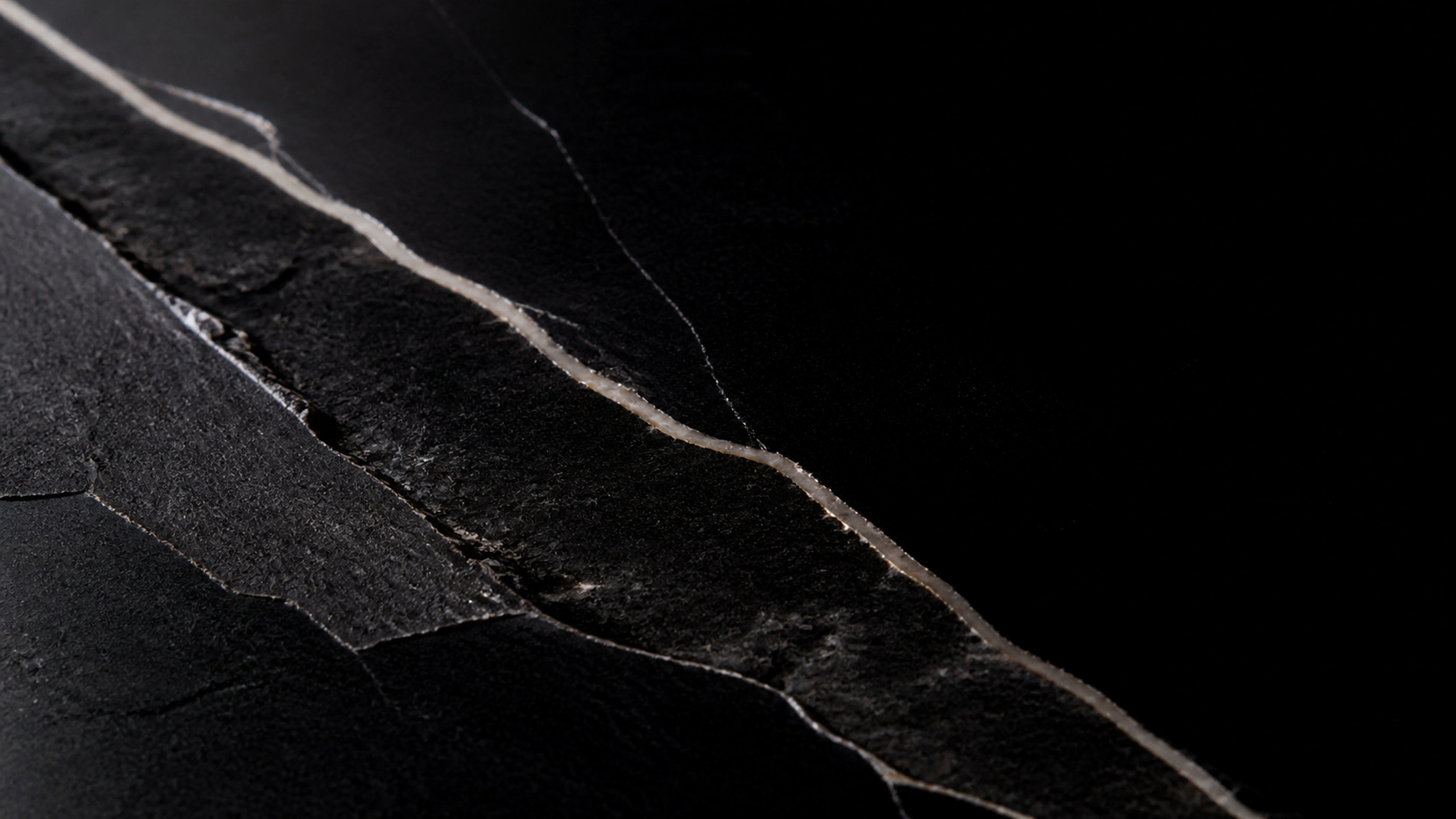 Extreme macro of rough black onyx split by one thin natural mineral vein, tactile stone detail fading into true black, original 4K dark amoled wallpaper