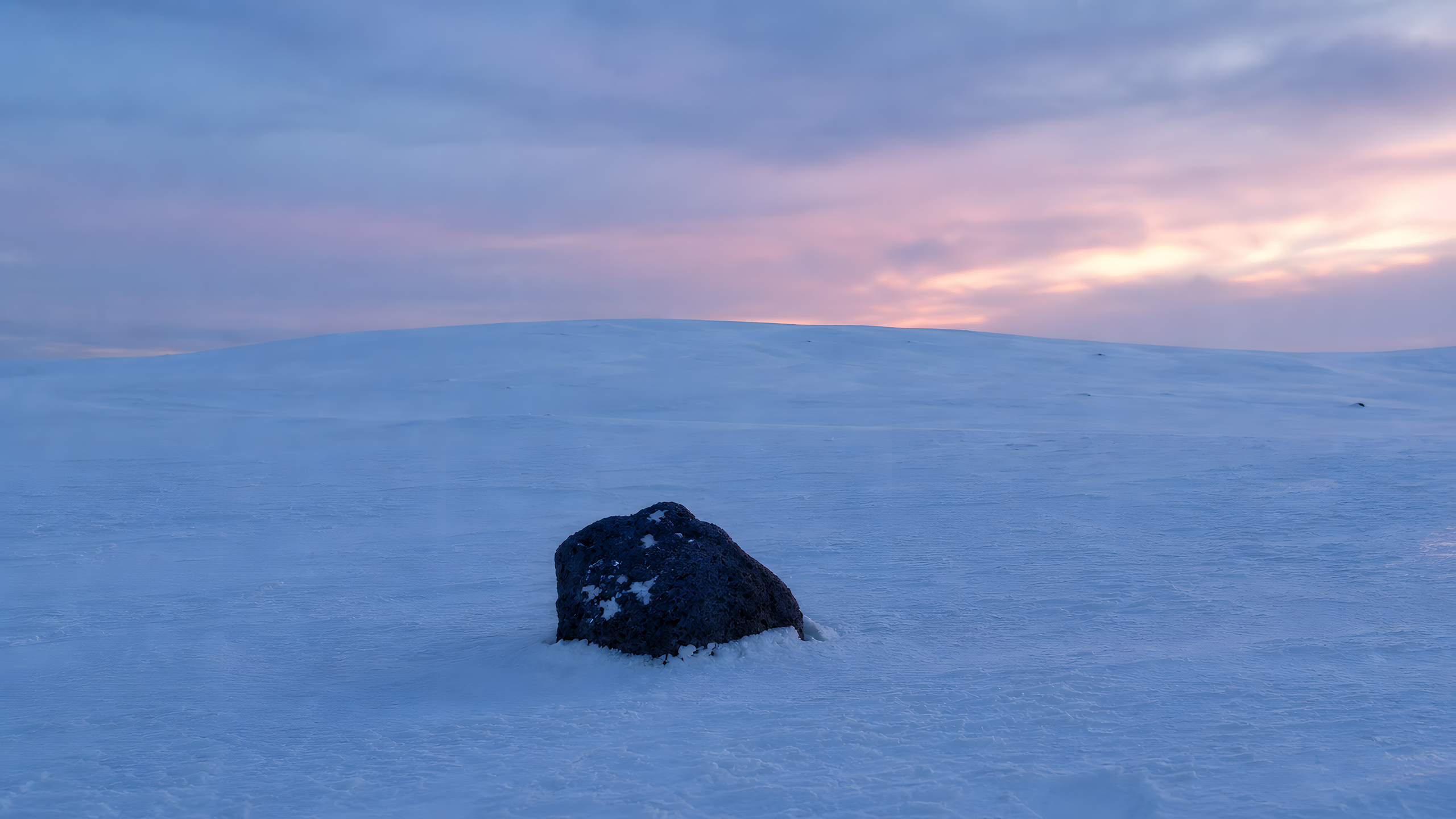 One weathered dark basalt boulder resting in an untracked winter snowfield beneath a distant ridge and quiet blue-hour afterglow, original 4K minimal wallpaper