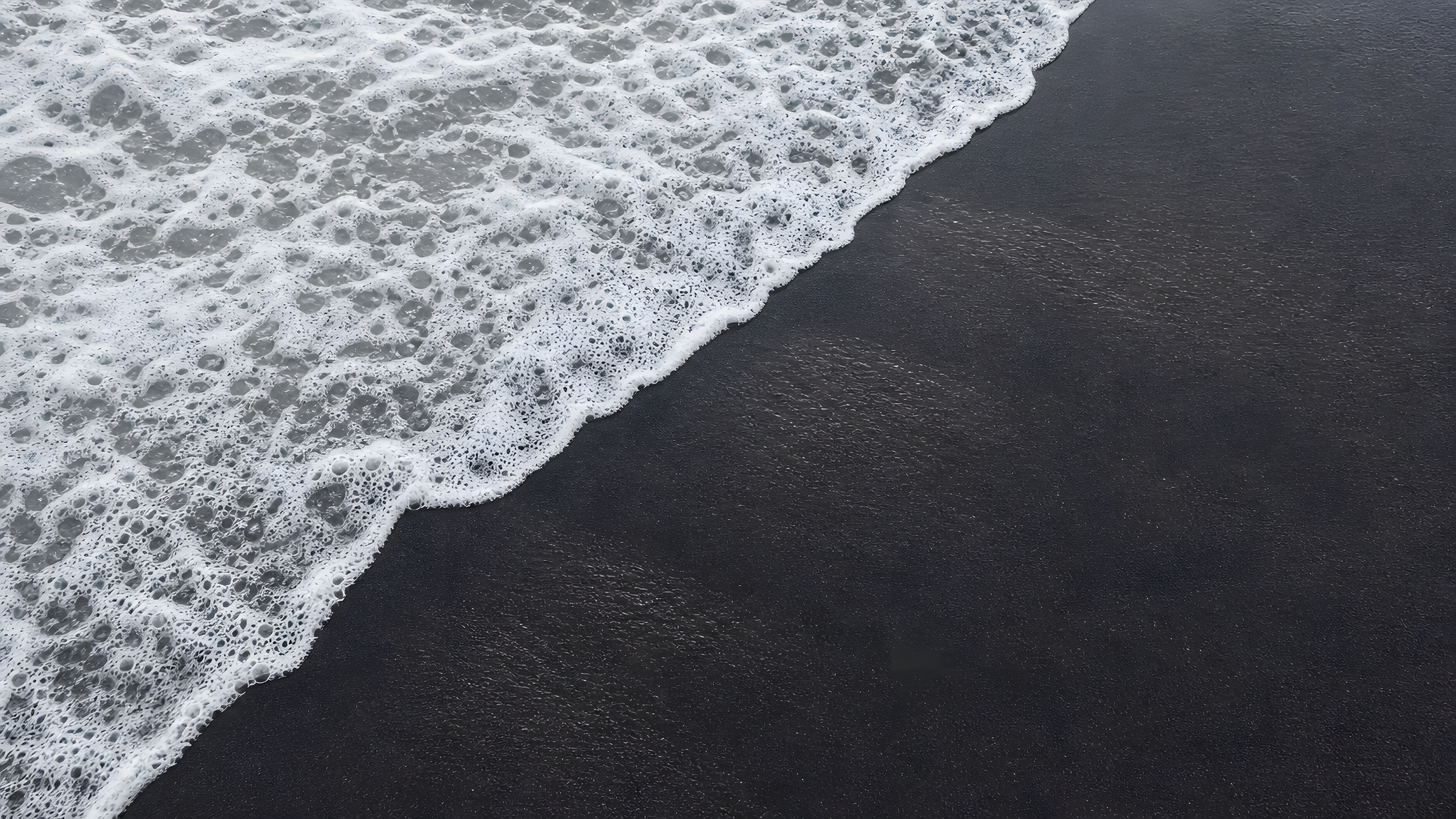 One clean ocean-foam curve retreating diagonally over smooth black volcanic sand in a true top-down view beneath bright overcast light, original 4K minimal wallpaper