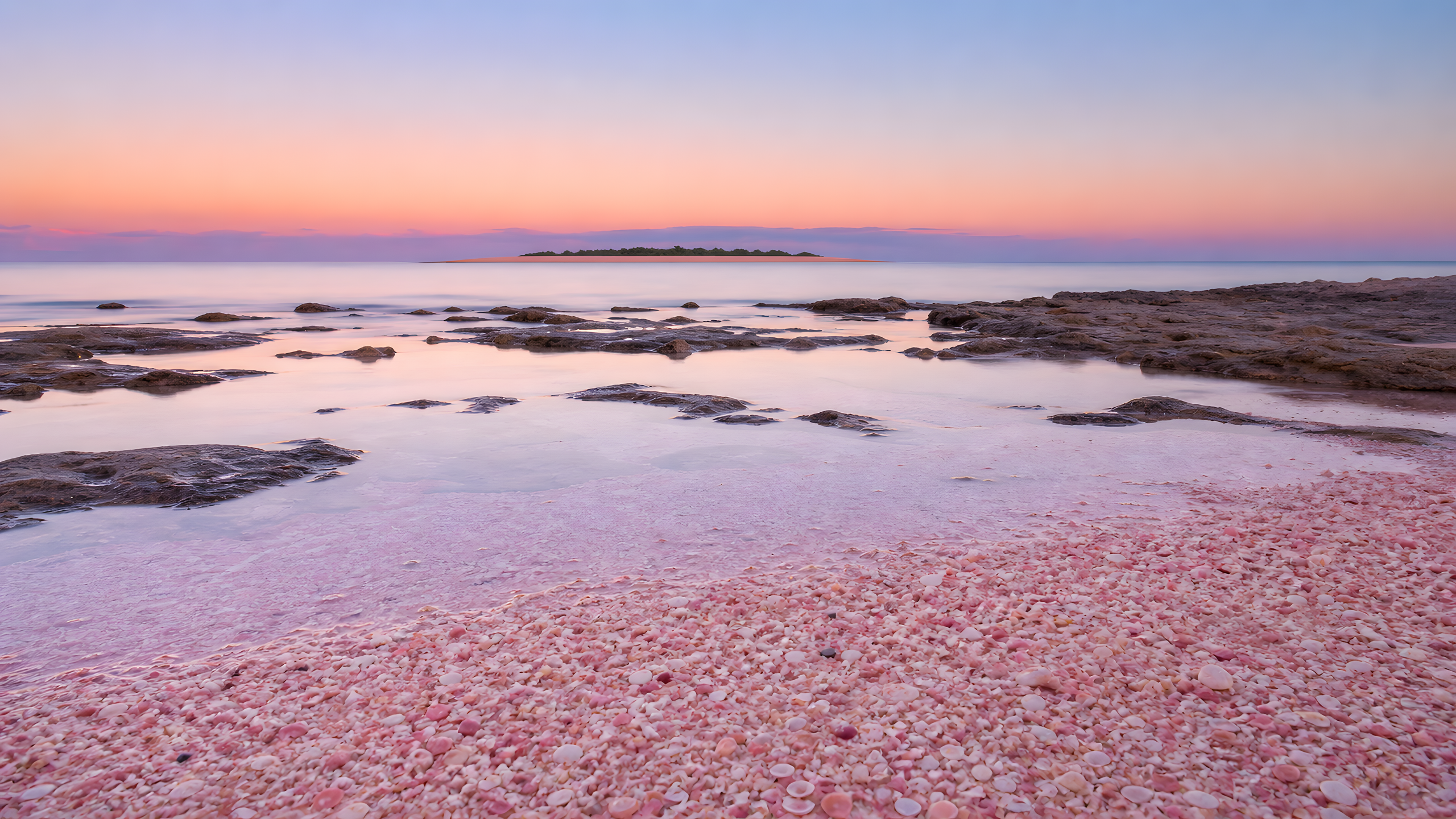 A quiet blush-pink crushed-shell beach with low coral shelves, glassy lavender water and a distant uninhabited cay beneath a peach-to-indigo dusk sky, original 4K beaches wallpaper