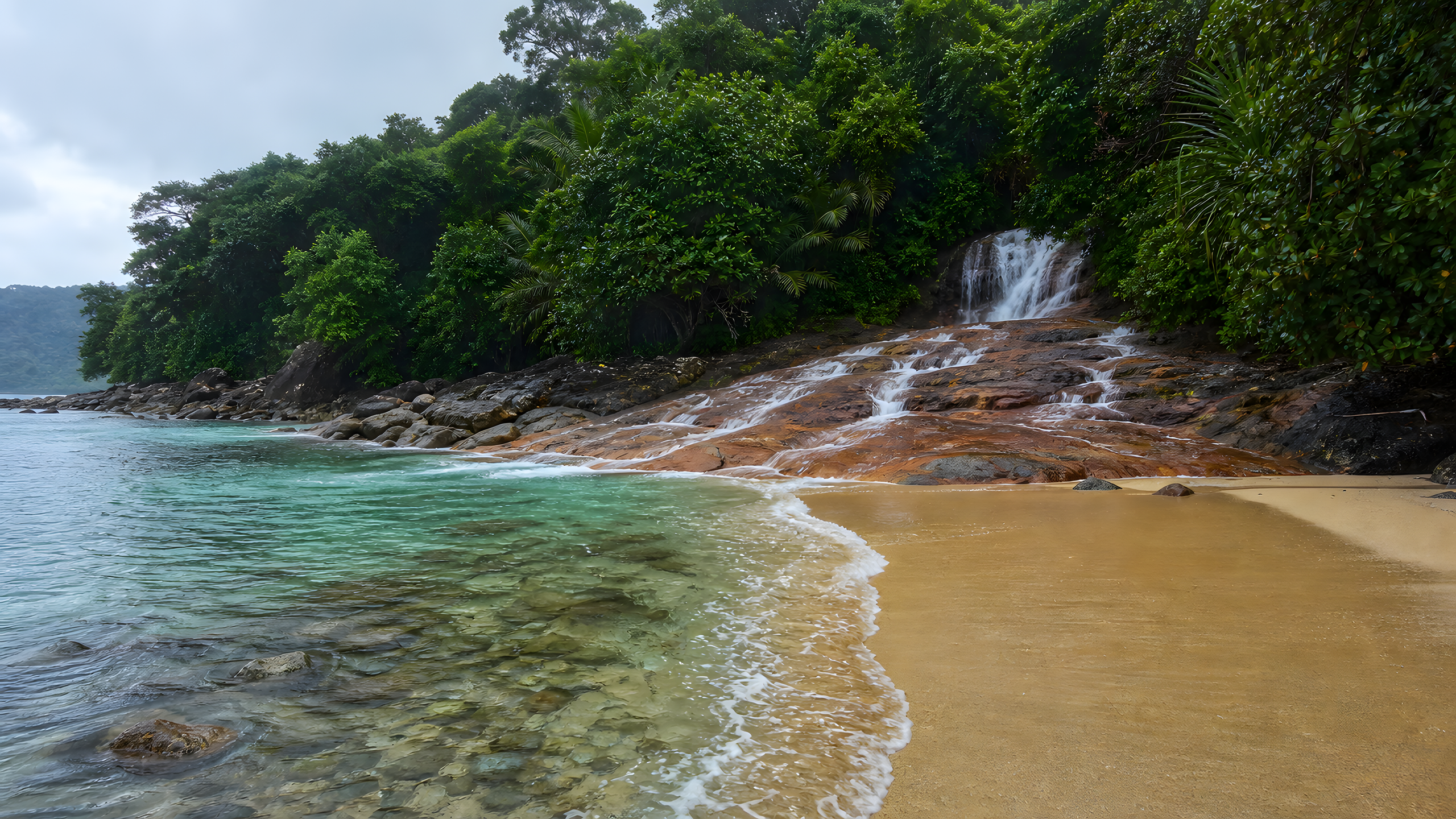 A freshwater cascade emerging from tropical rainforest and crossing a copper-stained rock shelf onto an empty golden beach beside clear teal sea, original 4K beaches wallpaper